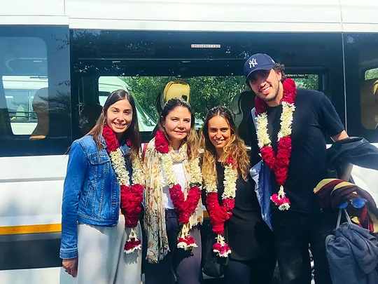 Cuatro viajeros sonríen con guirnaldas de flores rojas y blancas, junto a la van blanca, a su llegada a la India