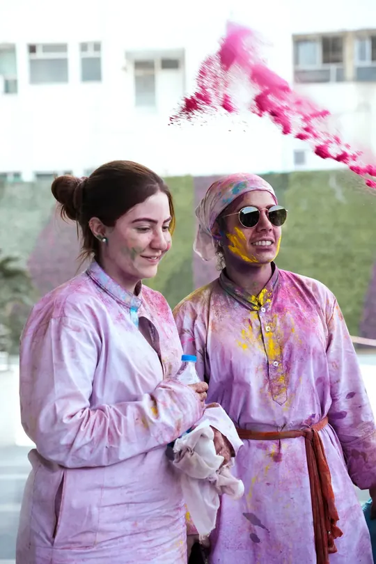 Dos viajeros vestidos de blanco y manchados de color sonríen mientras estalla una nube de polvo rosa sobre ellos durante el Holi